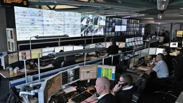 Mitarbeitende sitzen in zwei Reihen an Computerarbeitsplätzen mit fünf Bildschirmen. An der Wand vor ihnen sind große Monitore. Dort sieht man eine schematische Darstellung der Stadtbahnlinien und Bilder der Überwachungskameras an Haltestellen.