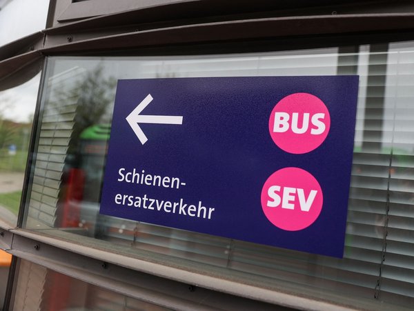 Blaues Schild auf Fenster: Weißer Pfeil nach links, darunter steht "Schienenersatzverkehr", rechts zwei pinke Kreise mit "BUS" und "SEV" 