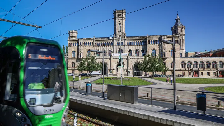 Links im Bild steht die ÜSTRA Straßenbahn, Linie 5 in Richtung Anderten, an der Haltestelle "Leibniz Universität". Im Hintergrund sind Gleise, die Universität, Menschen und Bäume zu sehen.