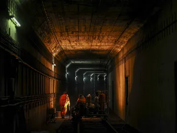 Zu sehen ist eine Lore und zwei Bauarbeiter in einem Stadtbahntunnel. Der Tunnel ist relativ dunkel, wird aber von einem warmen Licht erleuchtet. 