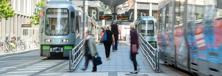 Menschen gehen die Rampe an einem Hochbahnsteig hinauf. An den Gleisen links und rechts sieht man silberne Stadtbahnen. Die rechte Bahn fährt die Haltestelle gerade an.