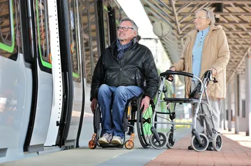 Ein Rollstuhfahrer und eine Dame mit Rollator stehen neben einer Stadtbahn des Typs TW 3000 an einem Bahnsteig und wollen einsteigen.