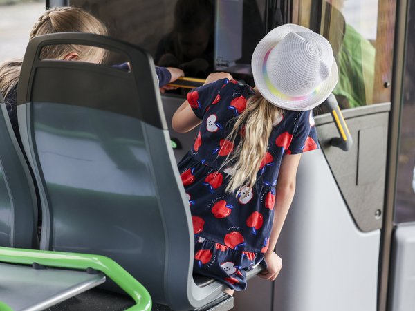 Blondes Mädchen mit Sommerhut von hinten. Sie sitzt im Bus, in der ersten Reihe hinter dem Busfahrer. 