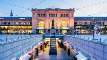 Panoramaaufnahme des Hauptbahnhofs Hannover bei Abendbeleuchtung, mit erleuchteter Fassade, Reiterstatue im Vordergrund und Treppenabgang zur Passerelle.