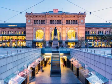Panoramaaufnahme des Hauptbahnhofs Hannover bei Abendbeleuchtung, mit erleuchteter Fassade, Reiterstatue im Vordergrund und Treppenabgang zur Passerelle.