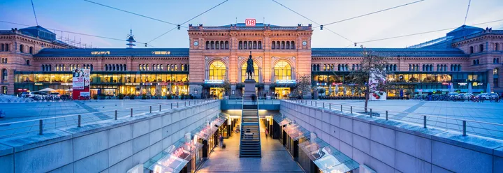 Panoramaaufnahme des Hauptbahnhofs Hannover bei Abendbeleuchtung, mit erleuchteter Fassade, Reiterstatue im Vordergrund und Treppenabgang zur Passerelle.