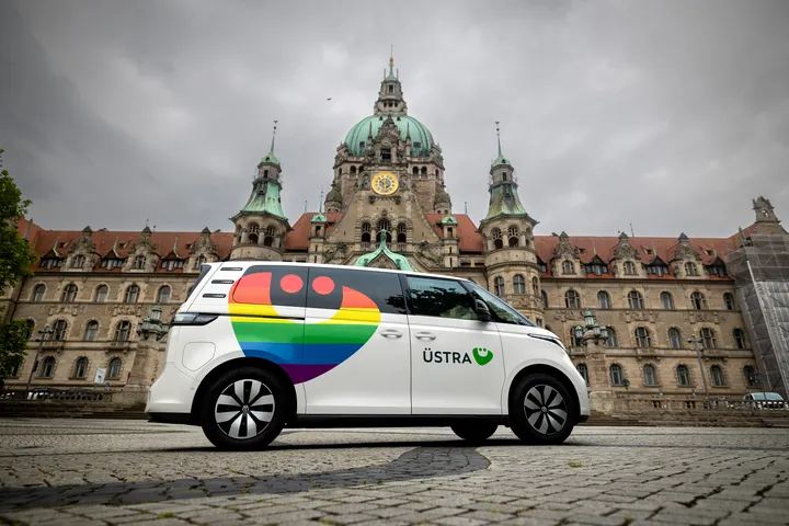 Zu sehen ist ein weißer ID Buzz vor dem Neuen Rathaus in Hannover. Das Fahrzeug ist mit einem bunten Regenbogen-ÜMO foliert und trägt das Logo der ÜSTRA. 
