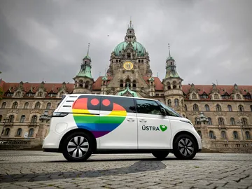 Zu sehen ist ein weißer ID Buzz vor dem Neuen Rathaus in Hannover. Das Fahrzeug ist mit einem bunten Regenbogen-ÜMO foliert und trägt das Logo der ÜSTRA. 