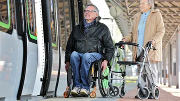 Ein Mann im Rollstuhl und eine Frau mit Rollator steigen in eine Stadtbahn. 