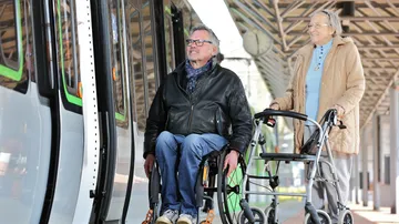 Ein Mann im Rollstuhl und eine Frau mit Rollator steigen in eine Stadtbahn. 