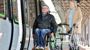 Ein Mann im Rollstuhl und eine Frau mit Rollator steigen in eine Stadtbahn. 
