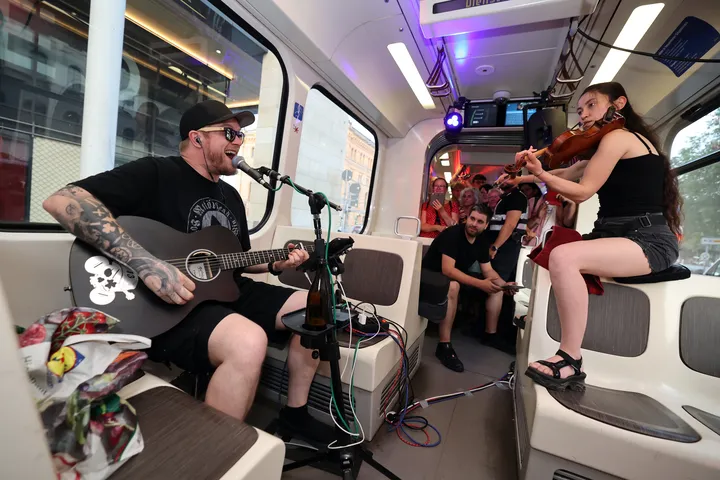 Man sieht zwei Musiker in einer Stadtbahn. Links sitzt Steven Screamz mit Gitarre und einem Mikrofon vor sich, rechts sitzt eine Geigerin. Im Hintergrund stehen Menschen, die zuhören.