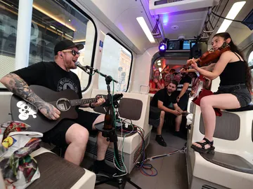 Man sieht zwei Musiker in einer Stadtbahn. Links sitzt Steven Screamz mit Gitarre und einem Mikrofon vor sich, rechts sitzt eine Geigerin. Im Hintergrund stehen Menschen, die zuhören. 