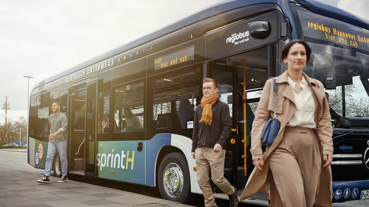 Ein eCitaro Bus mit sprintH Beklebung. Drei Fahrgäste verlassen den Bus: Ein Mensch an der hinteren Tür, zwei gehen durch die vordere Tür. Sie lächeln.