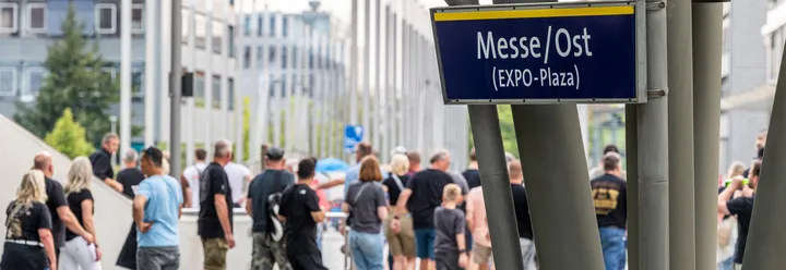 Es ist das Schild der ÜSTRA Haltestelle "Messe/Ost (EXPO-Plaza)" zu sehen. Im Hintergrund sind Menschen zu sehen, die verschwommen sind.