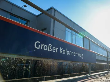 Zu sehen ist ein Haltestellenschild auf dem "Großer Kolennenweg" steht. Die Schrift ist weiß, der Hintergrund des Schildes dunkelblau. Im Hintergrund sieht man ein Gebäde mit Flachdach, blauen Himmel und die Sonne. 
