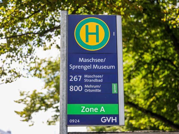 Zu sehen ist ein Bushhaltestellenschild. Die Station heißt "Maschsee/Sprengelmusuem" und es fahren die Linien 267 und 800. Im Hintergrund sind unscharfe Blätter eines Baumes zu sehen und heller Himmel. 