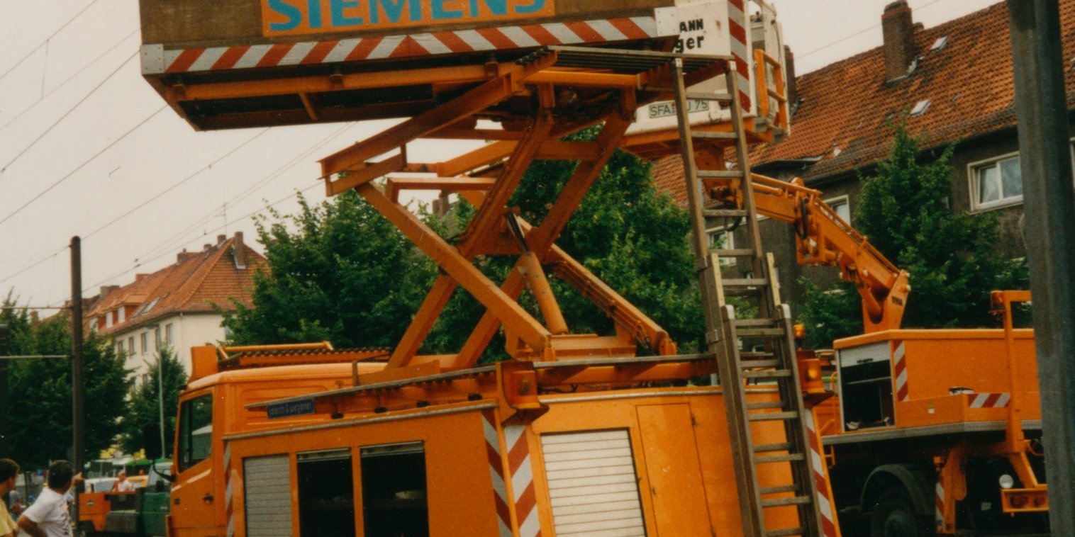 Ein Oberleitungswagen mit Hebebühne steht auf einem Gleis. Die Bühne ist angehoben. Drei Mitarbeitende stehen auf der Bühne. Ein Mitarbeiter steht auf den Mast der Oberleitung.