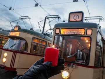 Eine Hand hält eine rote brennende Kerze, im Hintergrund sieht man mehrere historische Stadtbahnen mit der Fahrtzielanzeige "Friedenslicht".