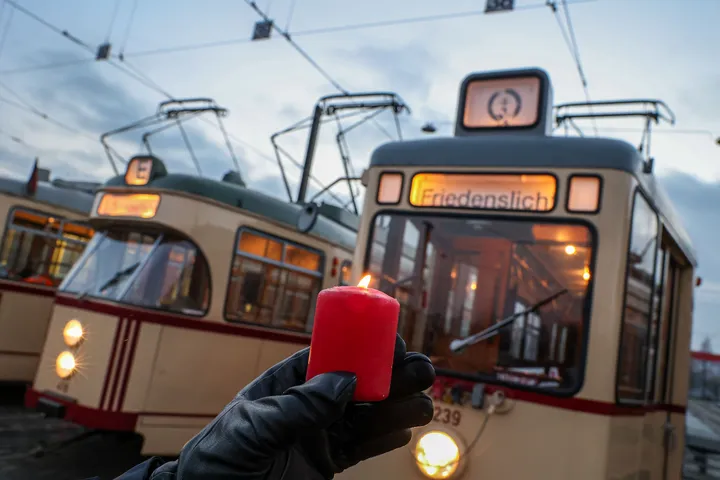 Eine Hand hält eine rote brennende Kerze, im Hintergrund sieht man mehrere historische Stadtbahnen mit der Fahrtzielanzeige "Friedenslicht".