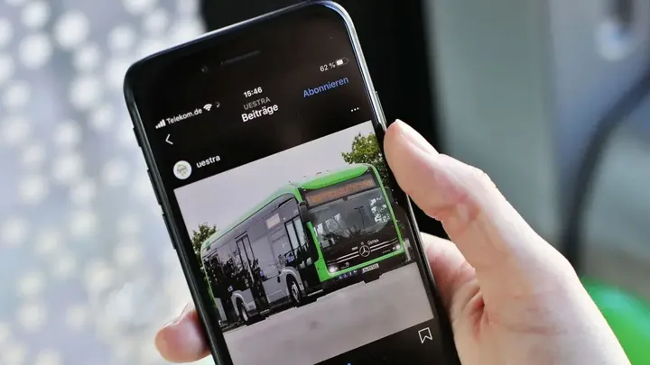 Eine Hand hält ein Handy. Das WLAN-Symbol ist aktiviert. Auf dem Bildschirm sieht man den Instagramkanal der ÜSTRA mit einem Foto eines eCitaro Busses. Im Hintergrund sieht man verschwommen das Sitzpolster und Fenster eines Busses.