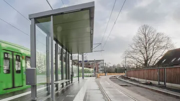 Der Hochbahnsteig mit Wartehäuschen der Haltestelle Riechersstraße im Regen. Ein TW 6000 fährt ein. 