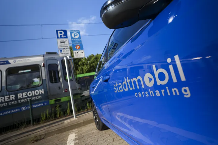 Seitliche Ansicht eines blauen Autos mit stadtmobil-Beklebung, das auf einem Parkplatz steht. Im Hintergrund fährt eine Stadtbahn des Typ TW2000 vorbei. 