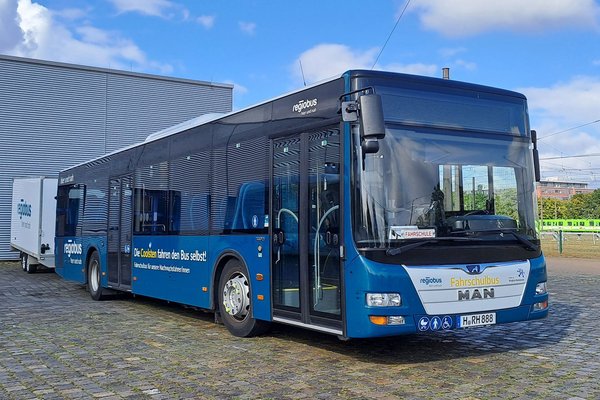 Ein MAN A20 mit petrolfarbener Beklebung und einem gelben Schriftzug an der Front "Fahrschulbus". Der Bus steht auf einem Betriebshof. Ein Fahrradanhänger ist an ihn gekuppelt.