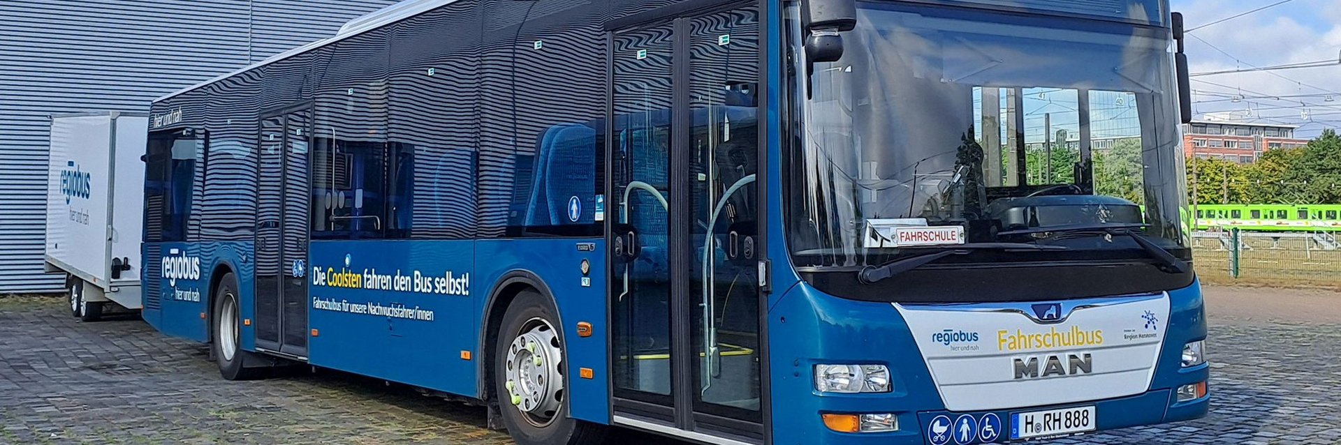 Ein MAN A20 mit petrolfarbener Beklebung und einem gelben Schriftzug an der Front "Fahrschulbus". Der Bus steht auf einem Betriebshof. Ein Fahrradanhänger ist an ihn gekuppelt.