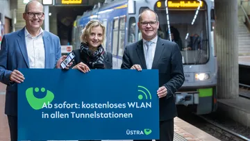 Ulf Birger Franz, Elke van Zadel und Grant Henning Tonne halten ein Schild mit dem Titel "Ab sofort: kostenloses WLAN in allen Tunnelstationen hoch.