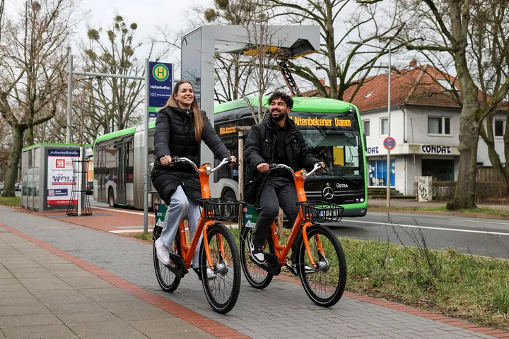 Zwei Radfahrer in der Innenstadt von Hannover auf orangenen Leihrädern von Donkey vor einem Lademasten und einem Bus 