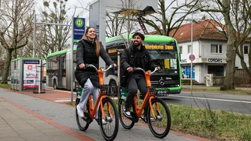Zwei Radfahrer in der Innenstadt von Hannover auf orangenen Leihrädern von Donkey vor einem Lademasten und einem Bus 