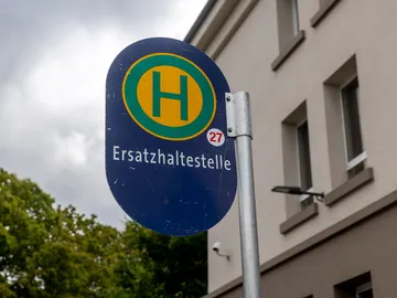 Im Fokus des Bildes ist das Schild einer Haltestelle zu sehen. Das Schild ist blau. Ein grünes großes H auf einem gelben Kreis. Darunter steht Ersatzhaltestelle. Im Hintergrund sieht man ein Stück Himmel mit grauen Wolken, rechts eine Baumkrone, links die Fassade eines Hauses.
