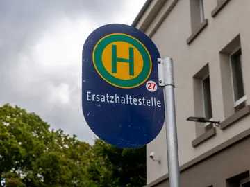 Im Fokus des Bildes ist das Schild einer Haltestelle zu sehen. Das Schild ist blau. Ein grünes großes H auf einem gelben Kreis. Darunter steht Ersatzhaltestelle. Im Hintergrund sieht man ein Stück Himmel mit grauen Wolken, rechts eine Baumkrone, links die Fassade eines Hauses.
