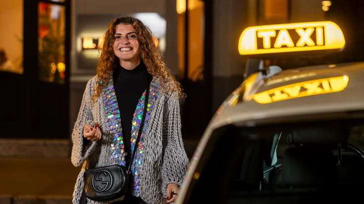 Eine Frau steht neben einem Taxi und schaut lächelnd in die Kamera. Das Taxi-Symbol leuchtet, im Hintergrund sieht man Fenster eines Gebäudes, die ebenfalls beleuchtet sind.
