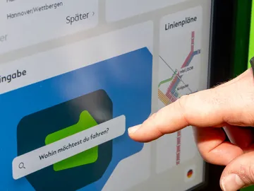 Eine Hand ist zu sehen, der Zeigefinger zeigt auf die Zieleingabe am Fahrkartenautomat der ÜSTRA. Hier steht: "Wohin möchtest du fahren?".