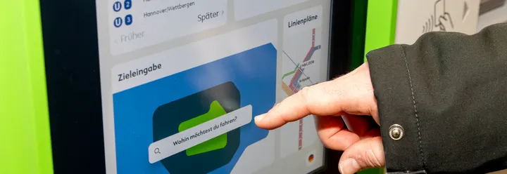 Eine Hand ist zu sehen, der Zeigefinger zeigt auf die Zieleingabe am Fahrkartenautomat der ÜSTRA. Hier steht: "Wohin möchtest du fahren?".
