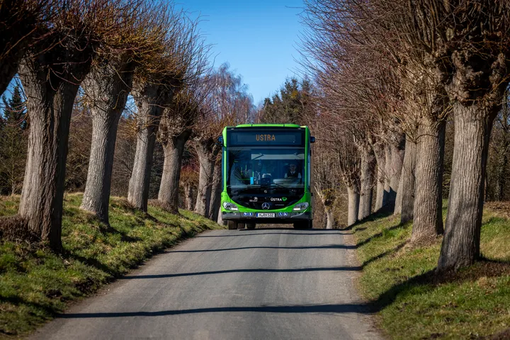 Ein ÜSTRA Bus fährt auf einem Weg zwischen Bäumen. Im Hintergrund sind Wiesen und Bäume zu sehen. 
