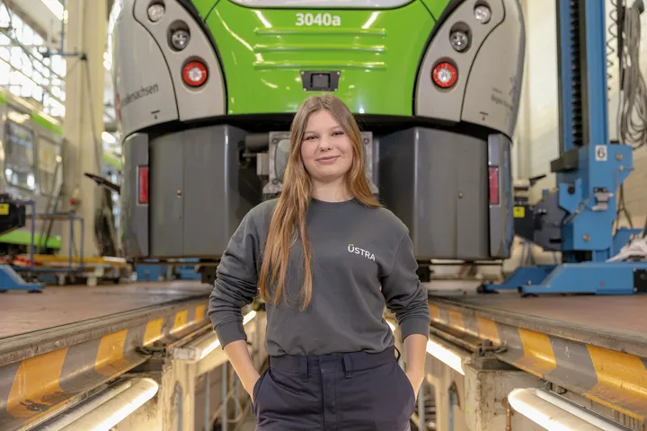 Eine junge Frau steht vor einer Stadtbahn in einer Werkstatt und schaut in die Kameras