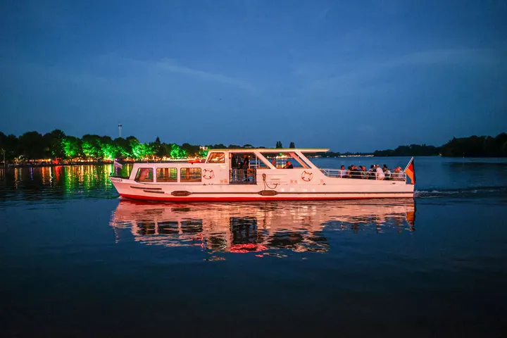 Ein Boot der Maschseeflotte fährt auf ruhiger See bei Dämmerung. Im Hintergrund ist bunt beleuchtetes Ufer.