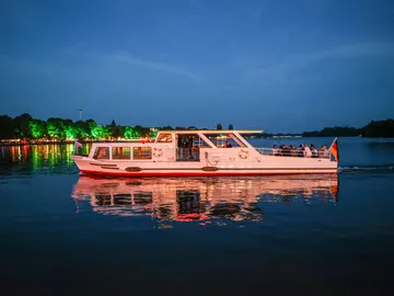 Ein Boot der Maschseeflotte fährt auf ruhiger See bei Dämmerung. Im Hintergrund ist bunt beleuchtetes Ufer.