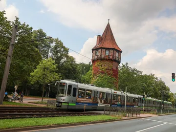 Im Hintergrund sieht man den Döhrener Turm in Hannover, aus roten Backsteinen mit spitzem Dach. Vor ihm fährt eine silberne Stadtbahn, ein TW2000 von rechts nach links durchs Bild. Im Vordergrund ist eine Straße zu sehen, im Hintergrund Bäume, etwas blauer Himmel und weiße und graue Wolken. 