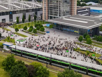 Eine Drohnenaufnahme zeigt eine ÜSTRA Stadtbahn an der Messe. Im Hintergrund sind viele Menschen, Bäume, das Messegelände und Gebäude zu sehen.