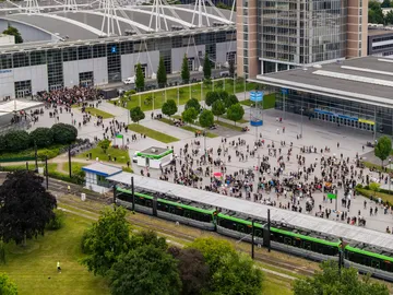 Eine Drohnenaufnahme zeigt eine ÜSTRA Stadtbahn an der Messe. Im Hintergrund sind viele Menschen, Bäume, das Messegelände und Gebäude zu sehen.