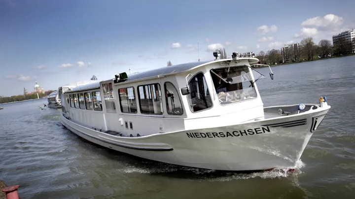 Das Schiff EMS Niedersachsen auf dem Maschsee in Hannover, im Hintergrund blauer Himmel, weiße Wolken und ein schmaler Streifen grüner Bäume. 
