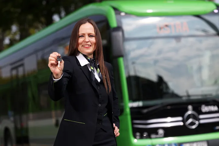 Das Bild zeigt eine ÜSTRA Fahrerin in Dienstkleidung vor einem Bus. Sie hält einen Schlüssel in der Hand. Es sieht aus, als würde sie jemandem den Schlüssel für den Bus anbieten.