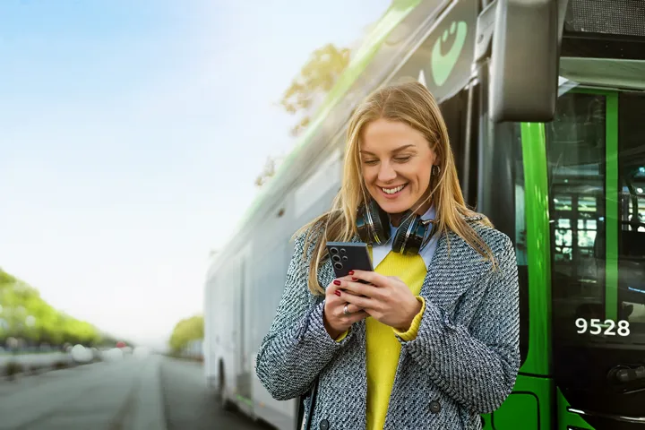 Frau steht vor einem Bus und schaut auf ihr Handy, dabei lacht sie