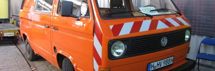 Ein VW Bus in Orange mit Warnleuchte und Reflektionselementen an der Front steht in einer Halle.