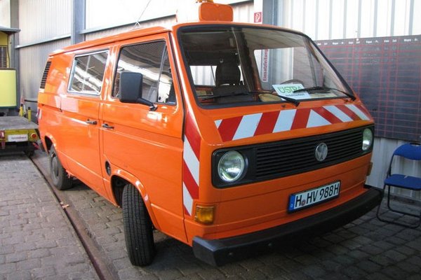 Ein VW Bus in Orange mit Warnleuchte und Reflektionselementen an der Front steht in einer Halle.