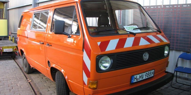 Ein VW Bus in Orange mit Warnleuchte und Reflektionselementen an der Front steht in einer Halle.